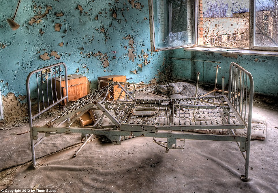 A broken bed-frame and bedside cabinets have been left by an open window, while once bright blue paint peels off the surrounding walls in a hospital ward. Shattered glass from the window litters the floor